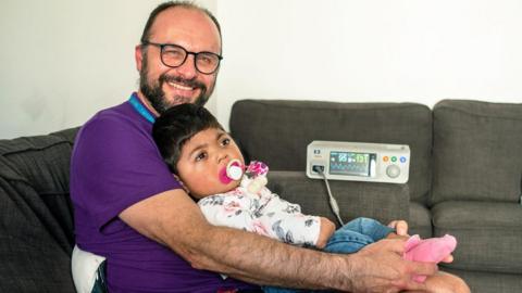 Aqsa's lying in the arms of her father Ikhlaq, she has a pink dummy in her mouth and pink fluffy slippers on her feet, while connected to a heart monitor. Her father is smiling at the camera, wearing a purple tee shirt and black glasses. 
