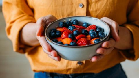A person in an orange shirt and denim trousers holds a bowl of vibrant berries in both hands.