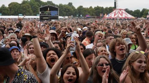 A crowd stands in front of a stage, some are clapping, others have their hands in the air and are singing along.