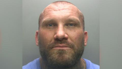 Mugshot of Derek Heggie, who is bald with a broad build and wearing a blue top. He has a short beard and moustache flecked with lighter coloured hairs.