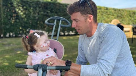 Harriet sits on her bike in the garden looking at her dad and smiling. He is wearing a grey top and smiles back at her. Harriet is wearing a pink outfit.