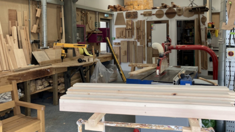 The image shows a woodworking shop filled with tools, materials, and equipment. There are stacks of wooden planks and boards on the left side.
A workbench with various tools. Several woodworking machines, including a table saw in the centre.