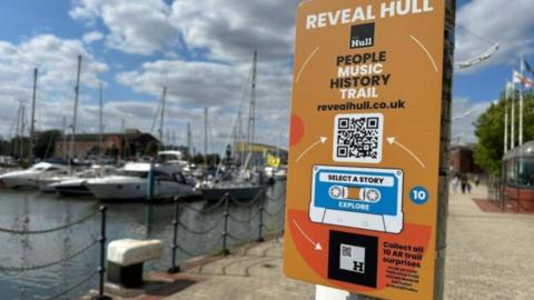 An orange sign with a blue and white cassette tape drawing on it. The writing says "Reveal Hull" above it and there is a QR code to access the immersive trail on the city's music history.