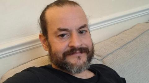 David Wragg with dark brown hair, beard and eyes wears a black t-shirt sitting on a beige sofa. 