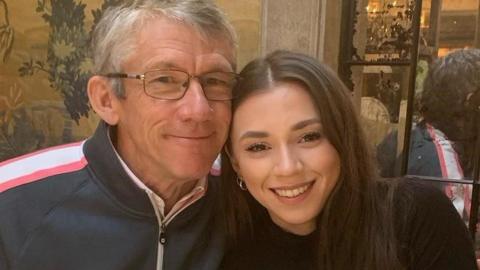 The image shows Peter Lawson and his daughter Thea Lawson, Peter is an older man with short grey hair and glasses, he's wearing a navy zip top with a white and stripe down the shoulder. His daughter is a young woman with long brown hair, she's wearing a black top. They sat close together with their heads touching.