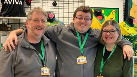 Members from the On The Stall City shop standing in a row looking directly at the camera and smiling. Andy Bowles is on the left dressed in a black T-shirt and grey zip up sweatshirt. He is wearing a pair of glasses and has a green lanyard round his neck. 