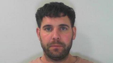 A man, in his forties, wearing a salmon-coloured top, with black hair and a short beard looks directly into the camera, stood in front of a white backdrop.