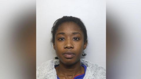 A woman in her twenties pictured in a police mugshot. She is wearing a grey fluffy jacket and a blue top underneath. She is staring directly at the camera.