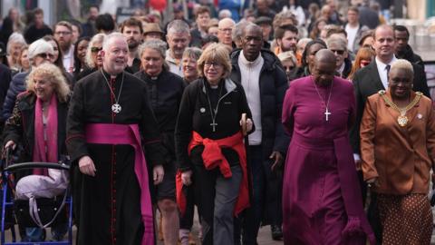 The Archbishop of Canterbury walking towards the camera with other religious leaders and 100 people behind her