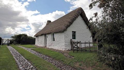 A small white building with a thatched roof and cobbled driveway and gate