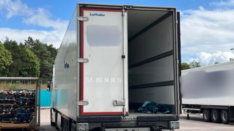 A large white lorry with one of its back doors open, showing sleeping bags on the bed of the lorry.