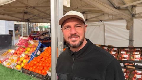 A man wearing a cap smiling, he is in a black quarter zip and is stood at a fruit stall. The stall has an assortment of brightly coloured fruit in under a white canopy