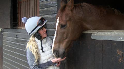A women wearing a jockey uniform pets a brown racehorse who is looking out from a stable.