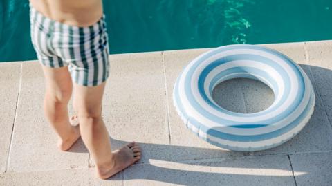 A child stood next to a swimming pool. They are wearing striped swimming trunks, and there is a rubber ring next to the pool.