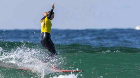 Lola Bleakley on a surfboard in the sea. Her arms are in the air.