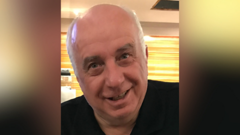 A headshot of a man smiling at the camera. He is wearing a black shirt. He has white hair around the sides of his head.