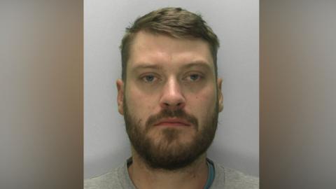 Custody image of Jordan Ireland. He is wearing a grey sweatshirt and has brown hair and a beard.