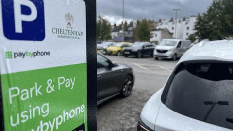 Cheltenham Borough Council parking metre in the forefront on the left hand side of the image. On the right is a close up of part of a car boot. Other cars in the car park are visible in the background.