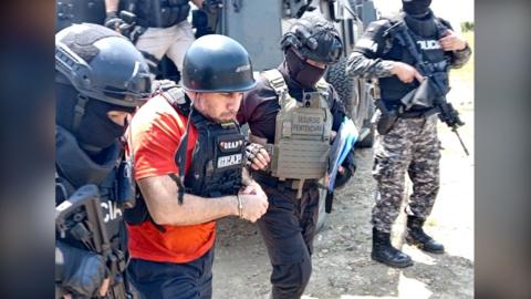 Ecuadorean drug lord 'Fito' wears a hard hat and an orange t-shirt underneath a bullet-proof vest. He is escorted by several people in black and camouflage clothes, also wearing bullet-proof vests.