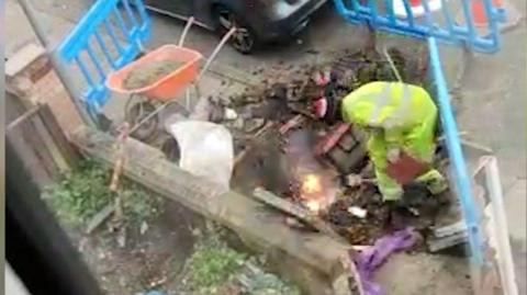 A man wearing yellow high-vis overalls and a woolly hat crouches over a hole in the pavement. He is surrounded by blue plastic barriers. A power cable is sparking and bursting into flames in front of him.