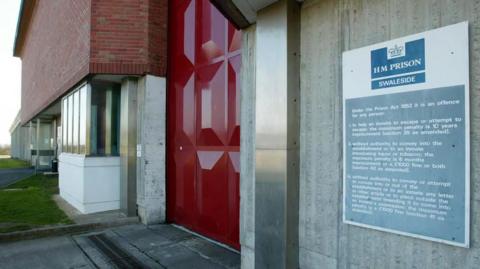 A close up image of a blue and white sign reading HM PRISON SWALESIDE, with smaller text below. It is stuck to a grey concrete wall. In the distance is a large red metal door.