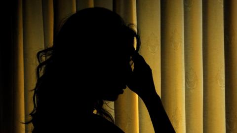 A silhouette of a woman holding her head. She looks sad and is standing in front of yellow curtains.