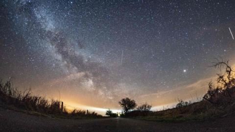 Masses of white stars shine out in the sky with the cloudy streak of the Milky Way from the left to the centre, with occasional white streaks in the sky from Lyrid meteors