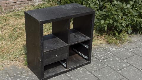 A black bookshelf with a draw is on a pavement outside a home. There is a brick wall and a green bush behind the unit.