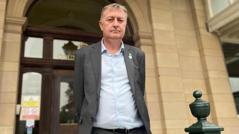 Man with grey hair in grey suit with light blue shirt stood outside a beige stone building with wooden door entrance
