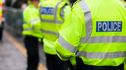 Three police officers wearing black trousers and yellow hi-vis jackets which say 'police' across the back in reflective silver letters. 