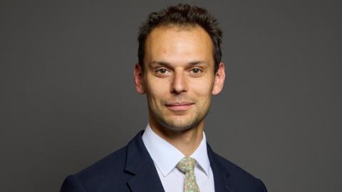 Josh Simons, a man with short, dark, curly hair, looks at the camera, wearing a navy jacket, white shirt and a pale green patterned tie 