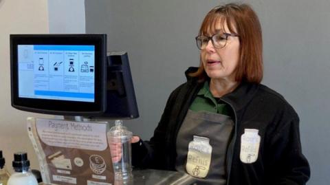 Rose King from Rose's Refills in her shop in Alsager.  She has dark red hair to her shoulders and wears round-rimmed glasses. She has a black fleece on over a grey apron which is over a green shirt. 