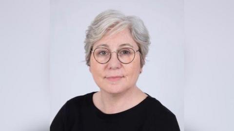 A head and shoulders photo of a woman in a black top with glasses and white hair. 