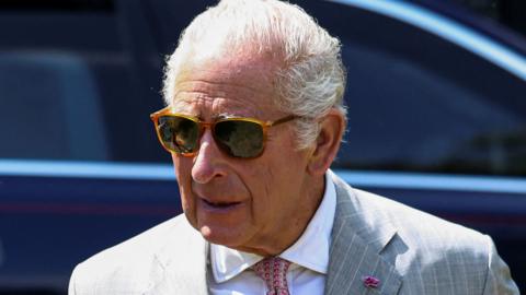 King Charles III in sunglasses and a grey suit walks away from a car