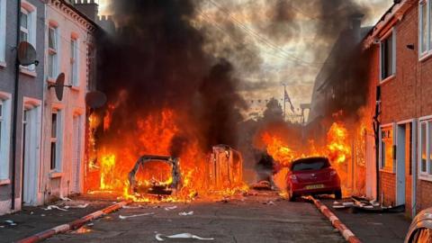 Cars and terrace homes on fire as black smoke rises into sky.