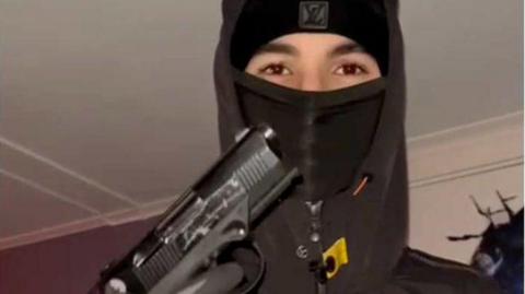 A teenager wearing a black balaclava and hoodie holding up a handgun, with his gloved hand on the trigger