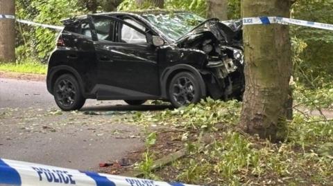 A black car has crashed into a tree. The bonnet of the car is caved in and the driver's window is covered by what appears to be an inflated airbag. There is police blue and white tape cordoning off the scene of the accident.