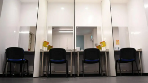 A general view of a safe drugs consumption room. Four chairs with black frames and a blue fabric are positioned in front of mirrors separated by white partitions. Yellow bins for sharps are attached to the walls. The floor is grey and reflections in the mirrors show a cupboard and a fire exit door.