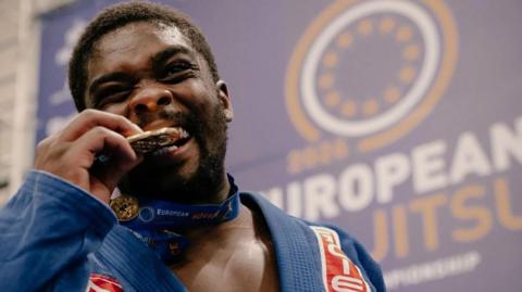 A man biting his medal. He is wearing a blue robe.