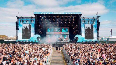 a packed crowd of people stand with their arms in the air in the sunshine below a music stage, decorated with giant blue wave and giant screens showing a performer on stage.