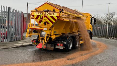 Yellow gritter dropping salt over road as it rides out of depot = SPREADING is in red letters on a yellow and red upside down triangle warning