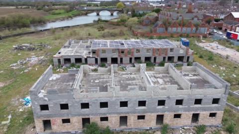 A drone view of two large, half-built apartment buildings standing on overgrown green land beside a wide river. The two-storey shells are open to the elements, with no door, windows or proper roofs. In the background stands a large Edwardian red-brick school with boarded-up windows.