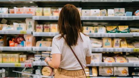 A woman in white shirt and mustard yellow trousers observes a fridge filled with cheese at a supermarket. She's holding a shopping basket in her left hand