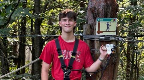 Hamish Wilson - a man wearing a red top and blue jeans, standing underneath a harness beside a tree.