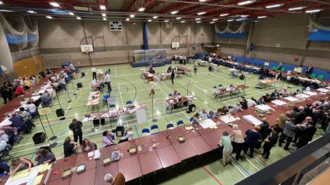 A sports hall with a green floor has a large number of brown tables set out in a horseshoe shape . There are lots of people counting ballot papers and lot of other people's looking closely. 