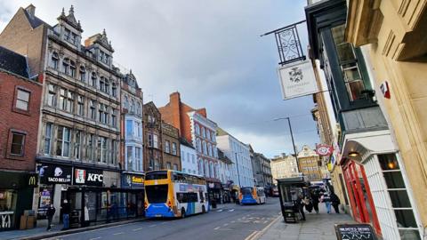 The Drapery in Northampton, with grand old buildings, and shops and restaurants at street level. There are buses that have pulled in at bus stops.