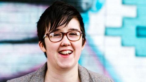 Lyra McKee, a woman with short black hair wearing glasses and a grey jacket, is smiling towards the camera. She is standing outside in front of a blue and pink mural on a wall.