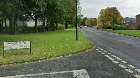 A sign for Earsdon Close is at the front of the image standing on a green lawn. It marks a junction with a much wider road, with trees and houses on either side