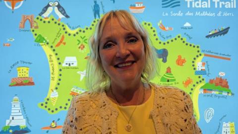 Tricia Warwick is pictured wearing a yellow dress and she has blonde hair. She is pictured in front of a map of Jersey that says tidal trail.