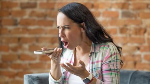 A woman with long black hair shouts into a phone she is holding in front of her. She is wearing a pink and green chequered shirt over a white t-shirt. Behind her is a brick wall.